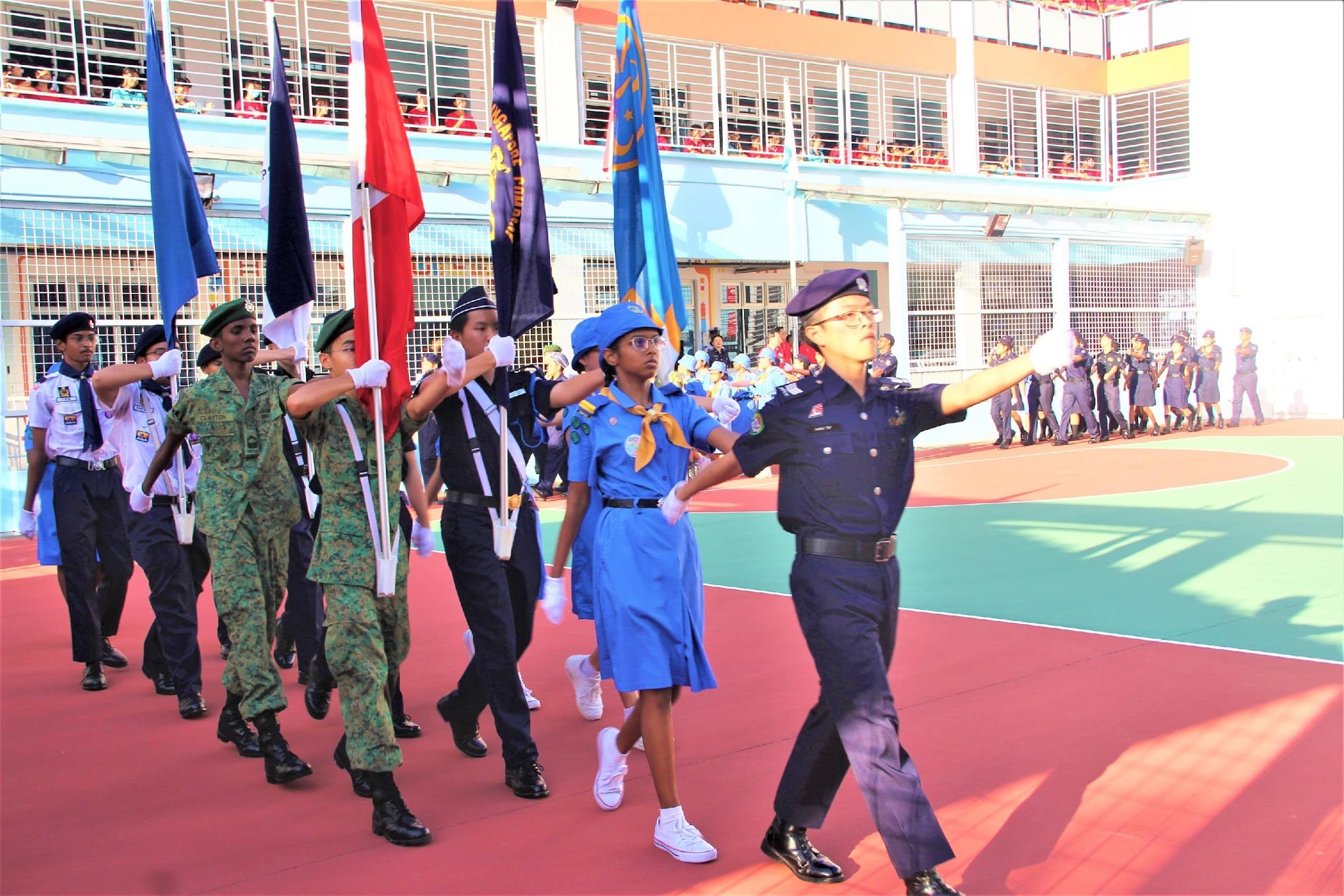 Students in various uniforms march in a parade carrying colorful flags on a red outdoor court.