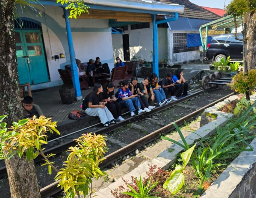 A group of young people sitting on railway tracks beside a white building with blue pillars and green plants nearby.