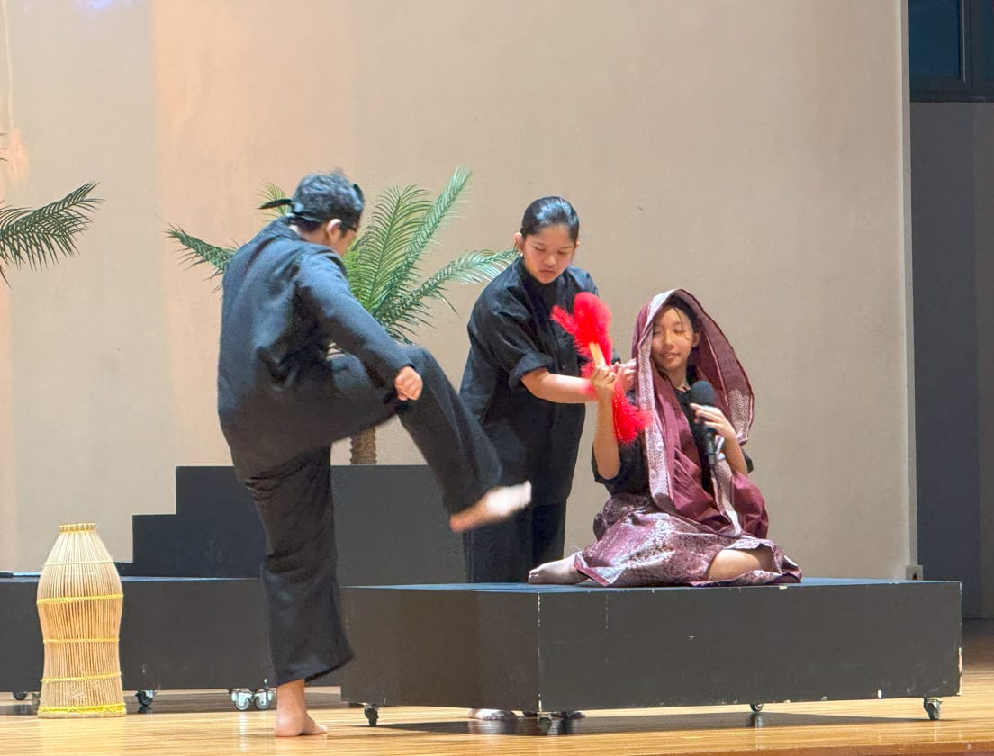 Three performers on stage, two in black costumes and one in purple, acting out a scene with a red fan.