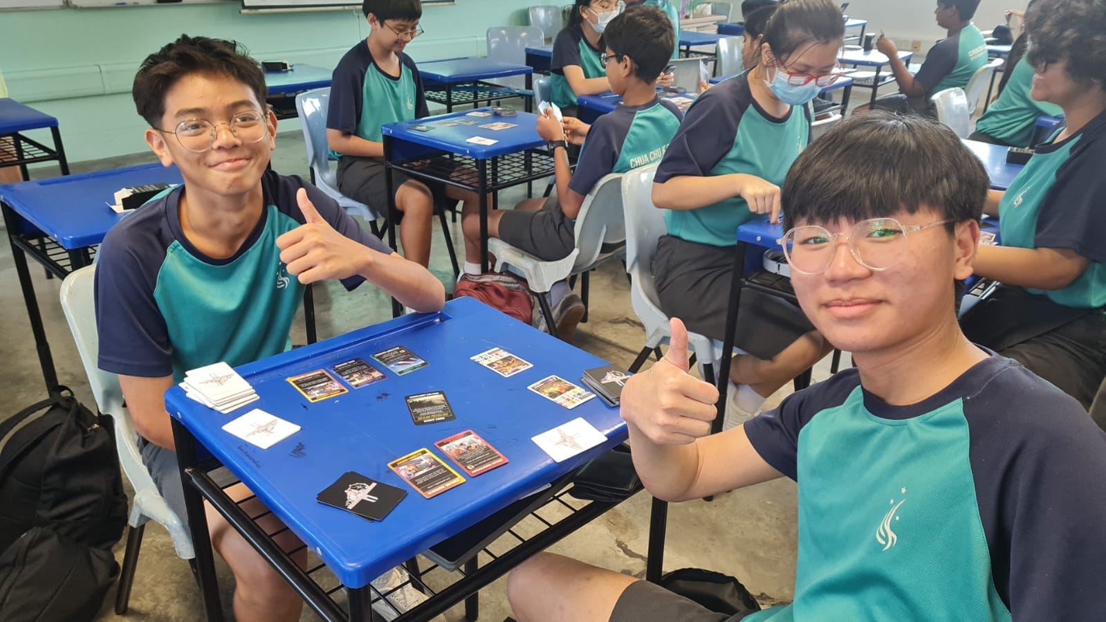 Two smiling students in teal uniforms give thumbs up across a blue desk with trading cards spread out in a classroom.
