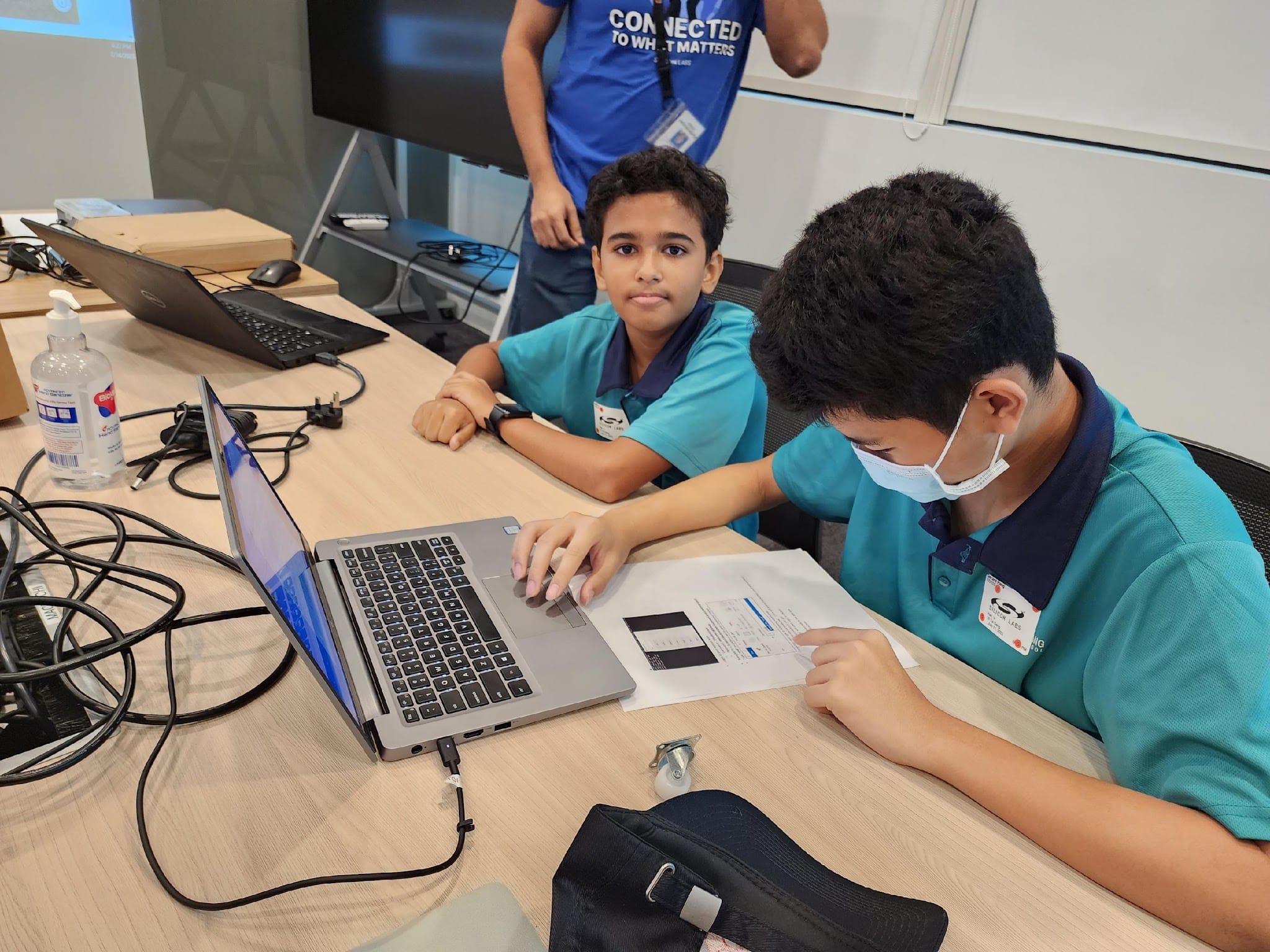 Two boys in teal uniforms work on a laptop at a desk, one wearing a face mask, with an adult standing behind.