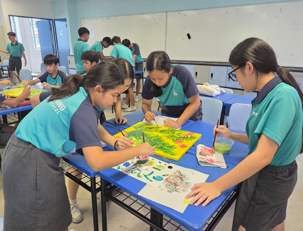 Students in teal uniforms paint colorful tropical leaf artwork on yellow canvas at blue desks in a classroom.