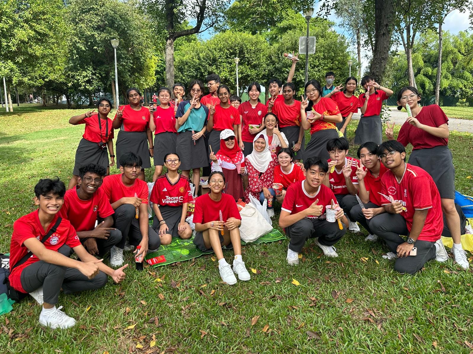 A large group of students mostly in red shirts pose and smile together on grass in a park surrounded by trees.