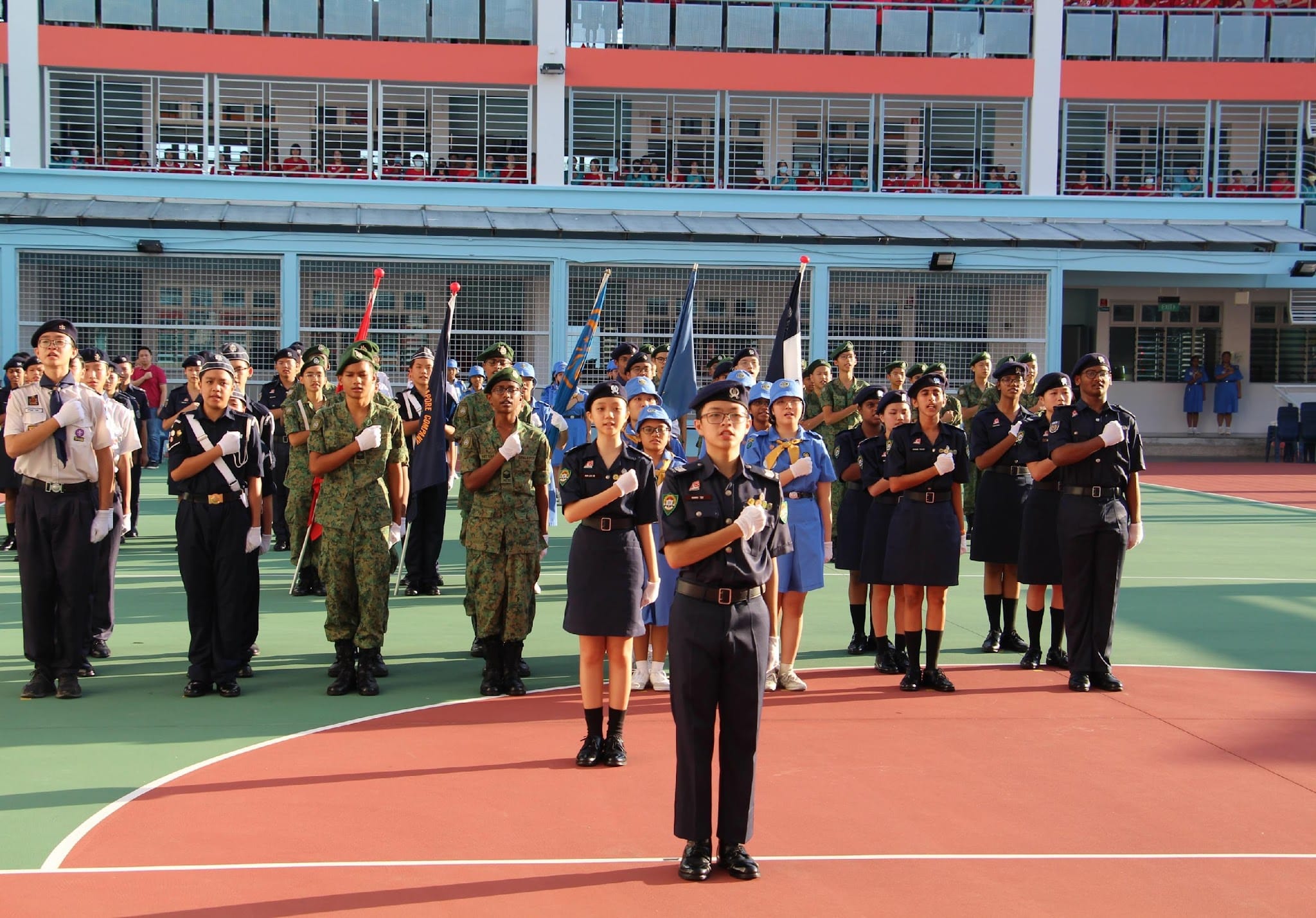 Students in various uniforms stand at attention during a formal parade on a school courtyard, with flags and spectators above.