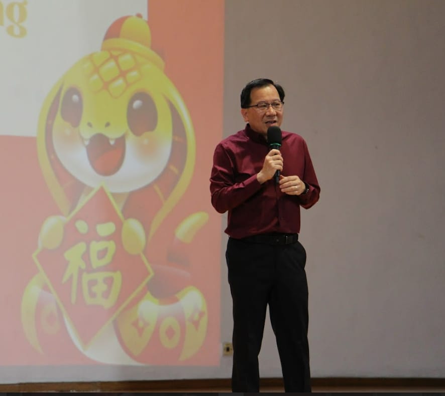 Man in dark red shirt holding a microphone speaks in front of a projected Chinese New Year snake cartoon slide.