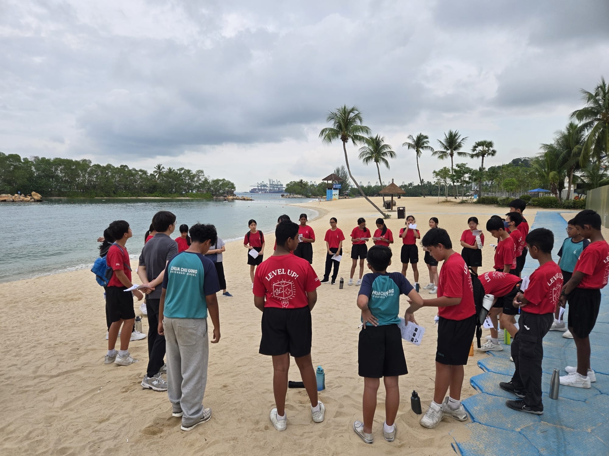 A group of students in red uniforms stand in a circle on a sandy beach with palm trees and calm water behind them.