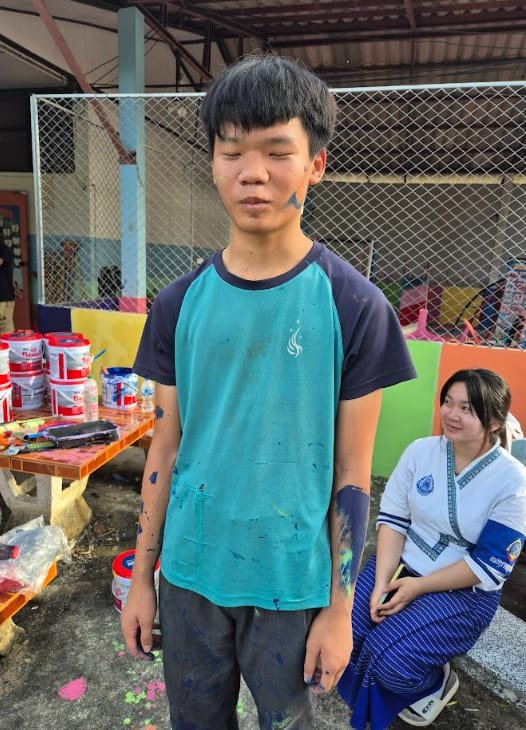 A teenage boy with blue paint on his face, arms, and teal shirt stands outdoors near paint buckets and colorful panels.