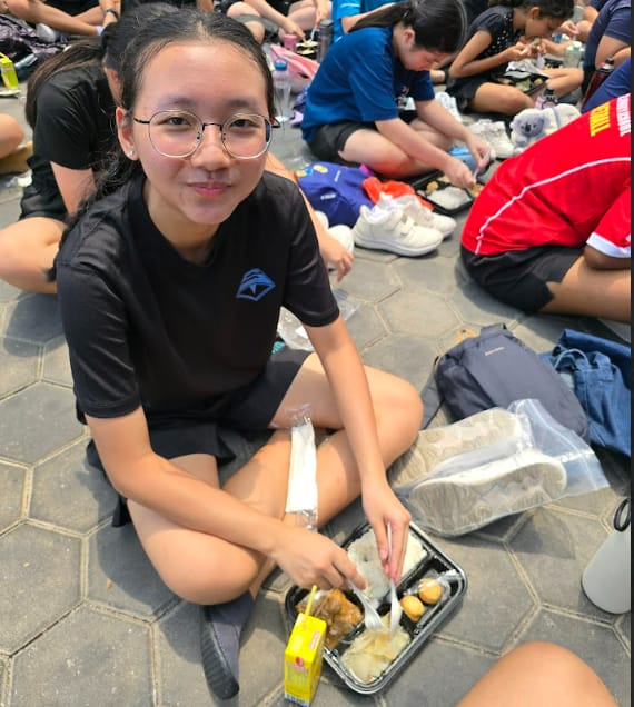 Young girl with glasses in a black shirt sits on pavement eating a meal from a tray, surrounded by other students.
