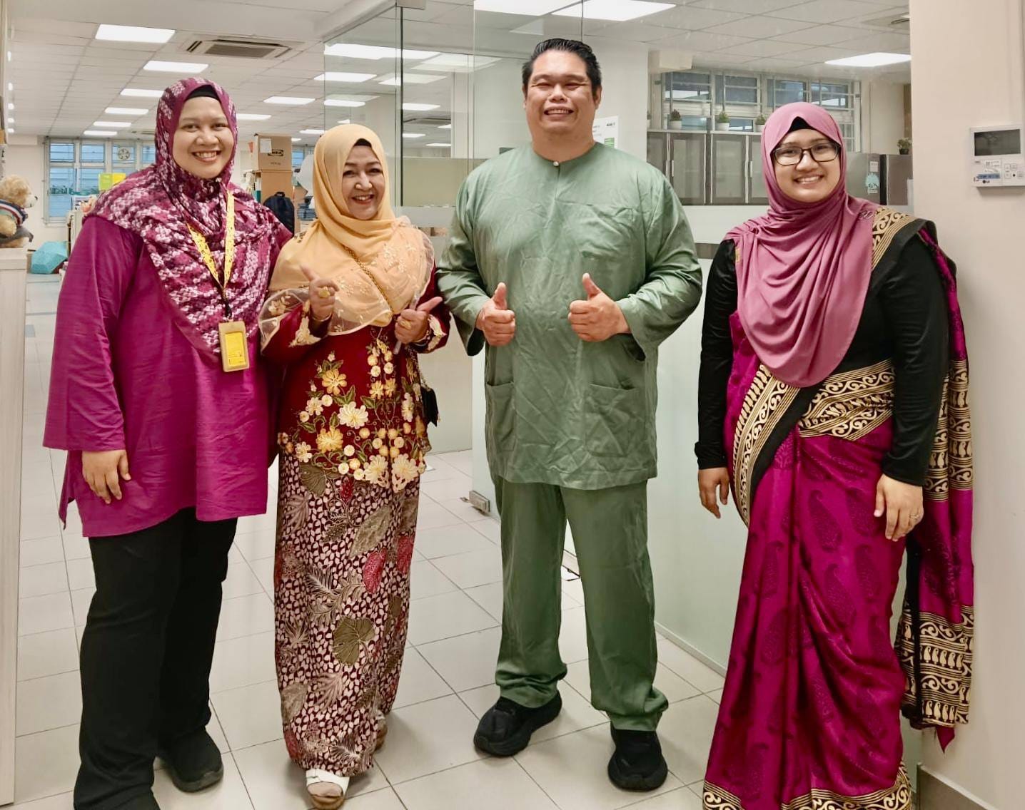 Four people in traditional Malay attire smile and give thumbs up inside an office setting.