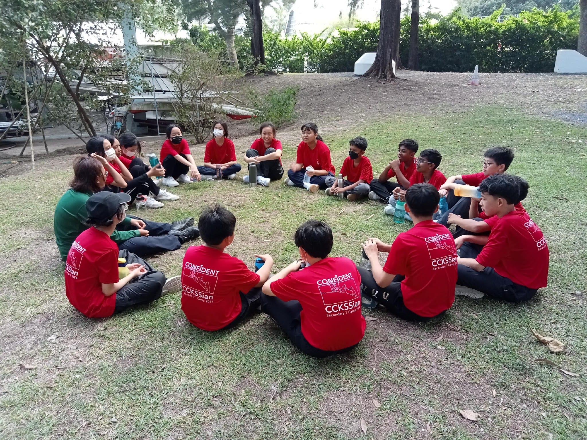 A group of students in red CCKSSian camp shirts sit in a circle on grass outdoors.