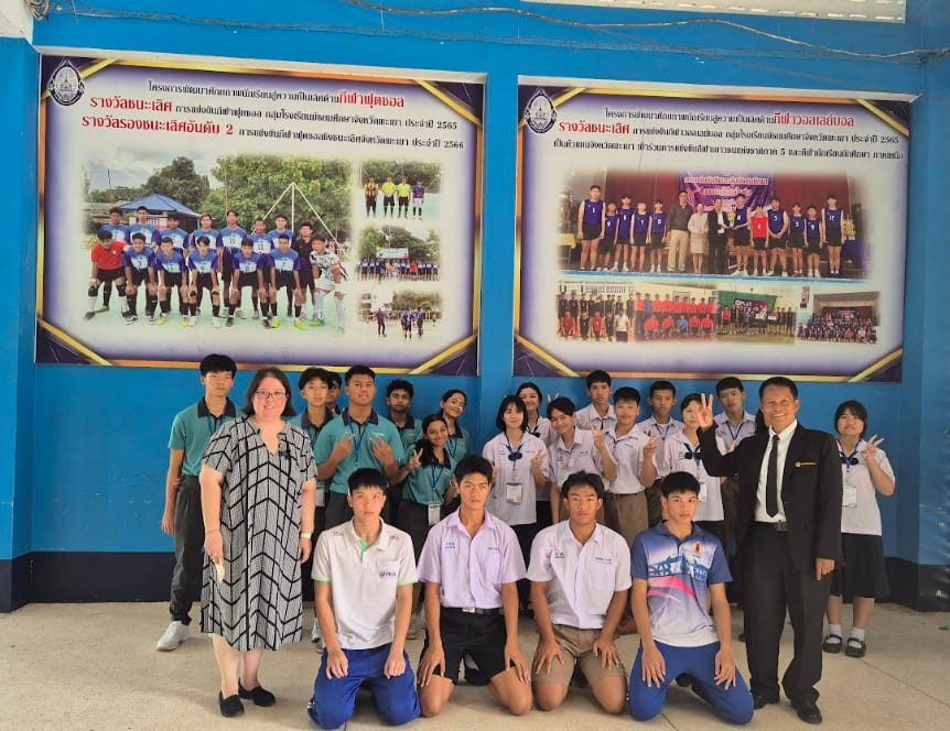 A group of students and teachers pose in front of two Thai sports award banners on a blue wall.