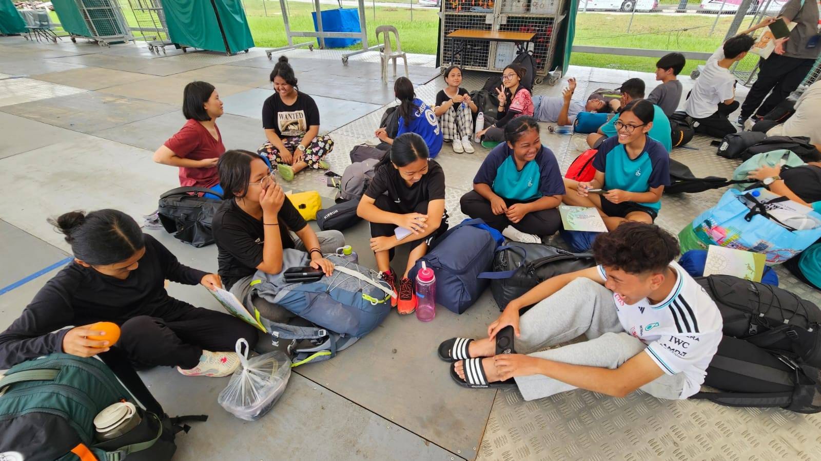 A group of young people sitting on a concrete floor outdoors with backpacks and bags, chatting and smiling.