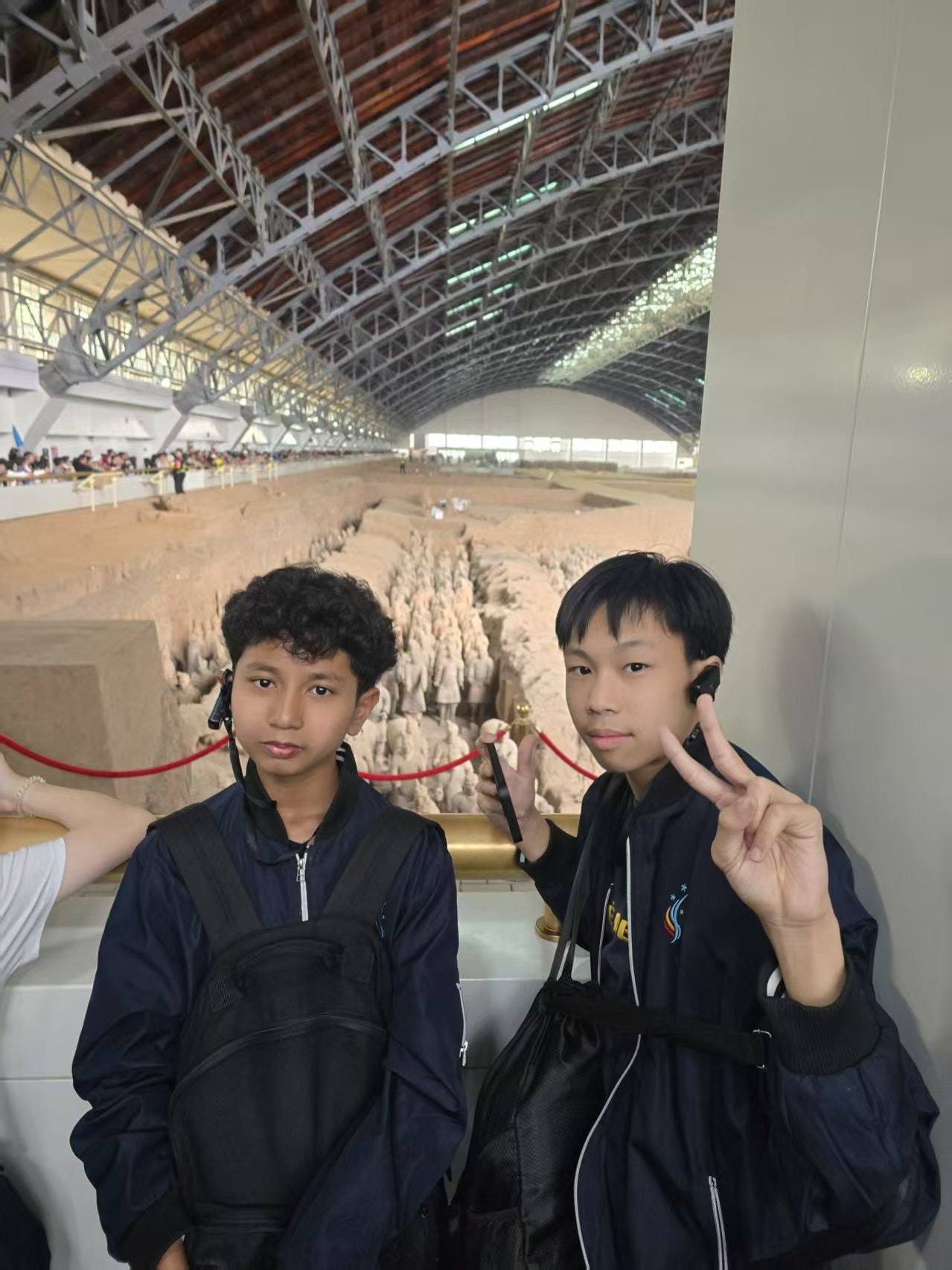 Two boys in navy jackets pose at the Terracotta Warriors museum, one making a peace sign, rows of clay soldiers visible behind them.