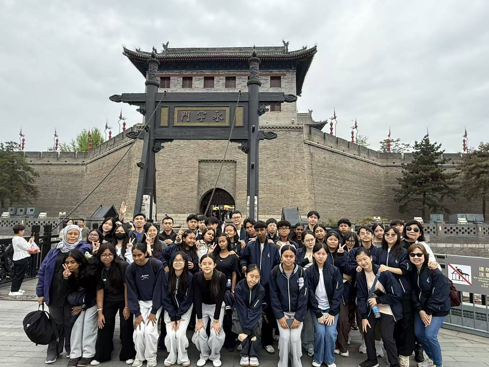 Large group of students and adults posing in front of a Chinese city wall gate with a drawbridge entrance.
