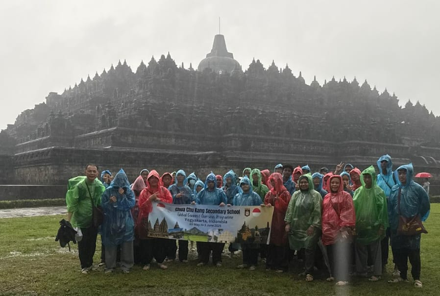 Group in colorful rain ponchos holds a Chua Chu Kang Secondary School banner in front of Borobudur temple.