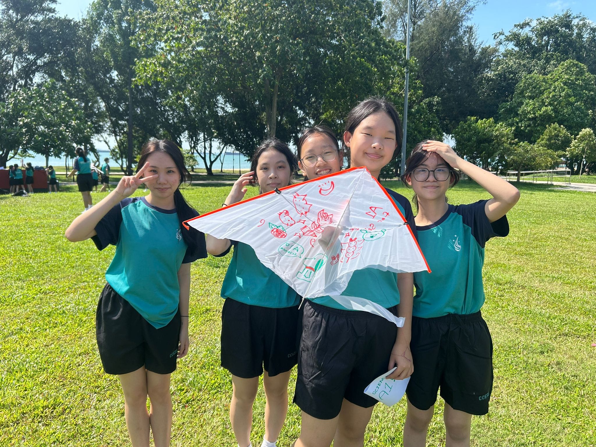 Five girls in matching teal uniforms stand on a grassy field, holding a hand-decorated white kite with red drawings.