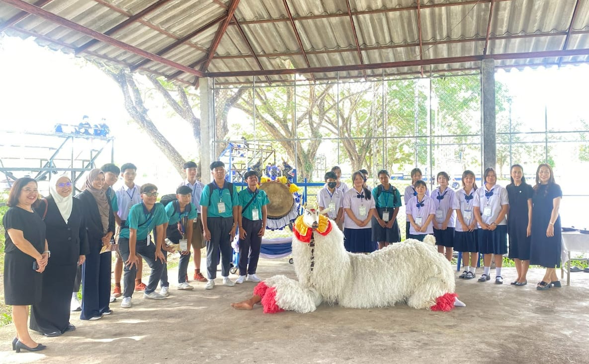 A group of students and adults pose under a shelter with a decorated white alpaca lying in the foreground.
