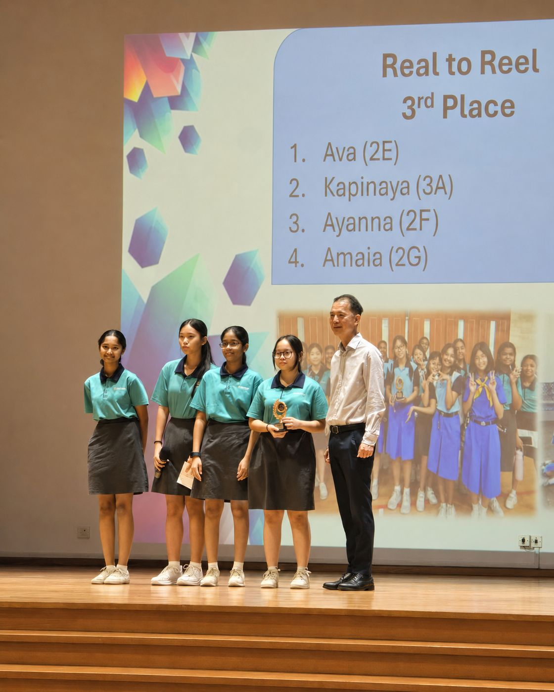 Four girls in teal uniforms stand on stage with a man beside a slide reading "Real to Reel 3rd Place."