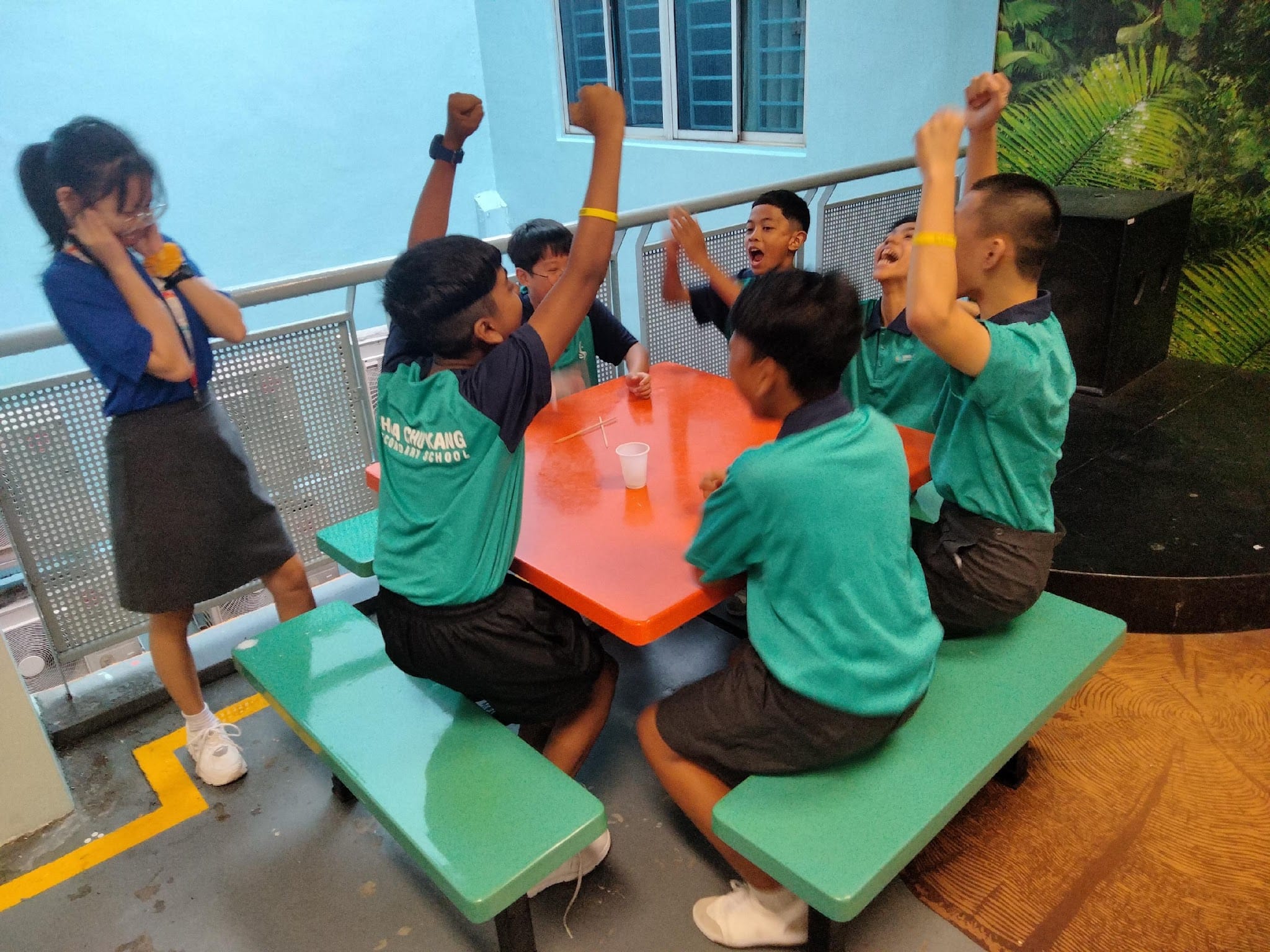 Several school students in green uniforms cheer with raised fists around an orange table with a plastic cup on it.