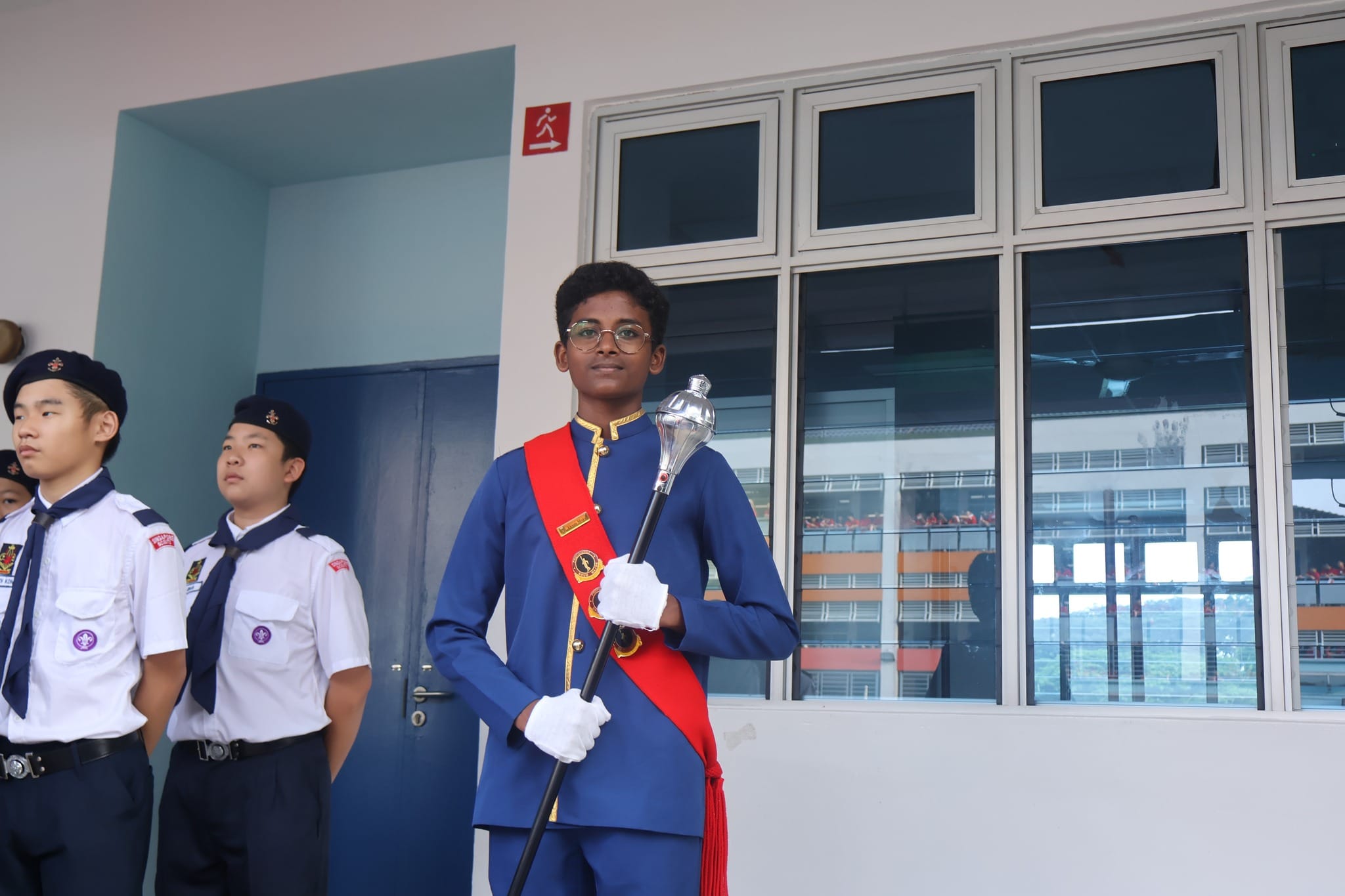 A young drum major in a blue uniform with a red sash holds a baton, with scouts in white uniforms standing behind.