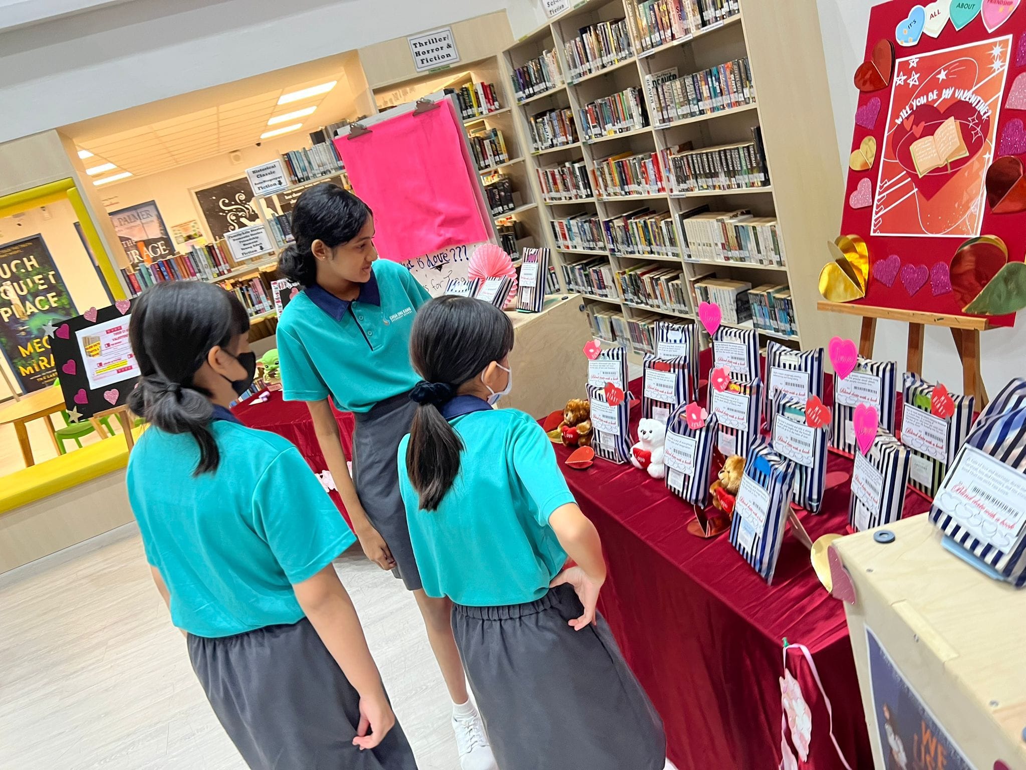 Three school girls in teal uniforms stand near a Valentine's Day display table with striped gift bags in a library.