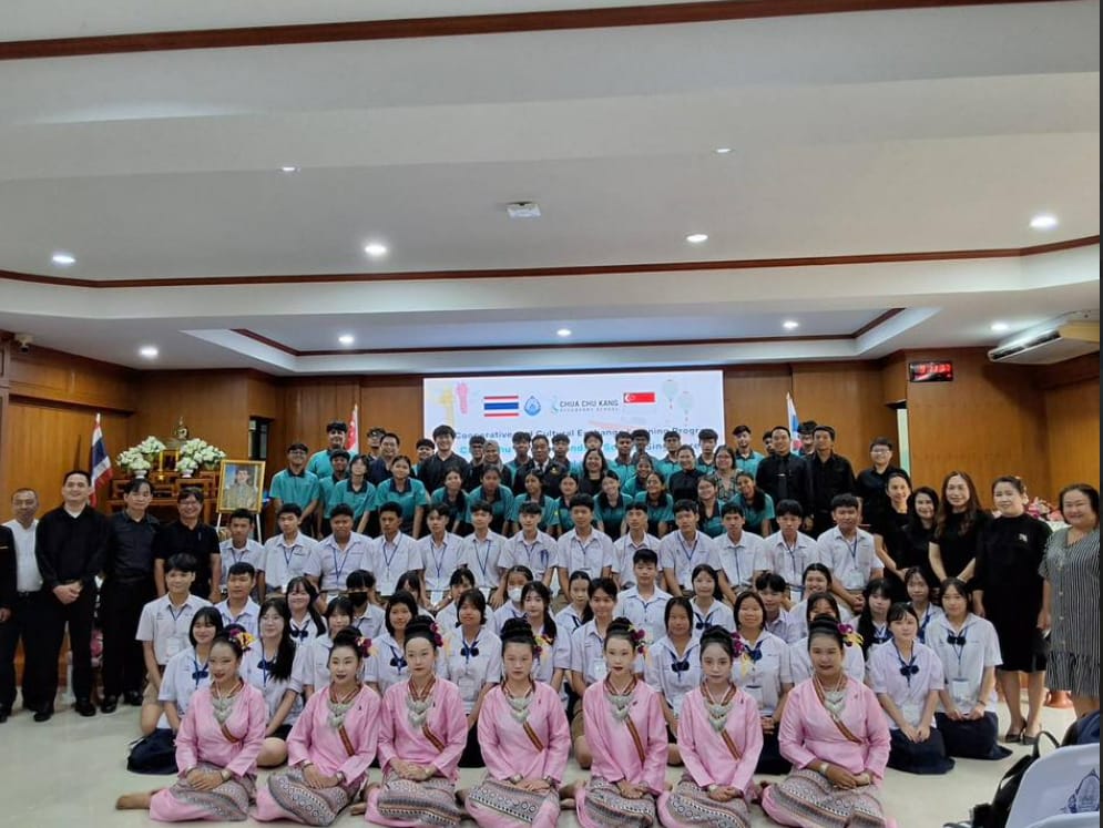 Large group of students and adults posing indoors for a cultural exchange program, with Thai and Singapore flags on a banner.