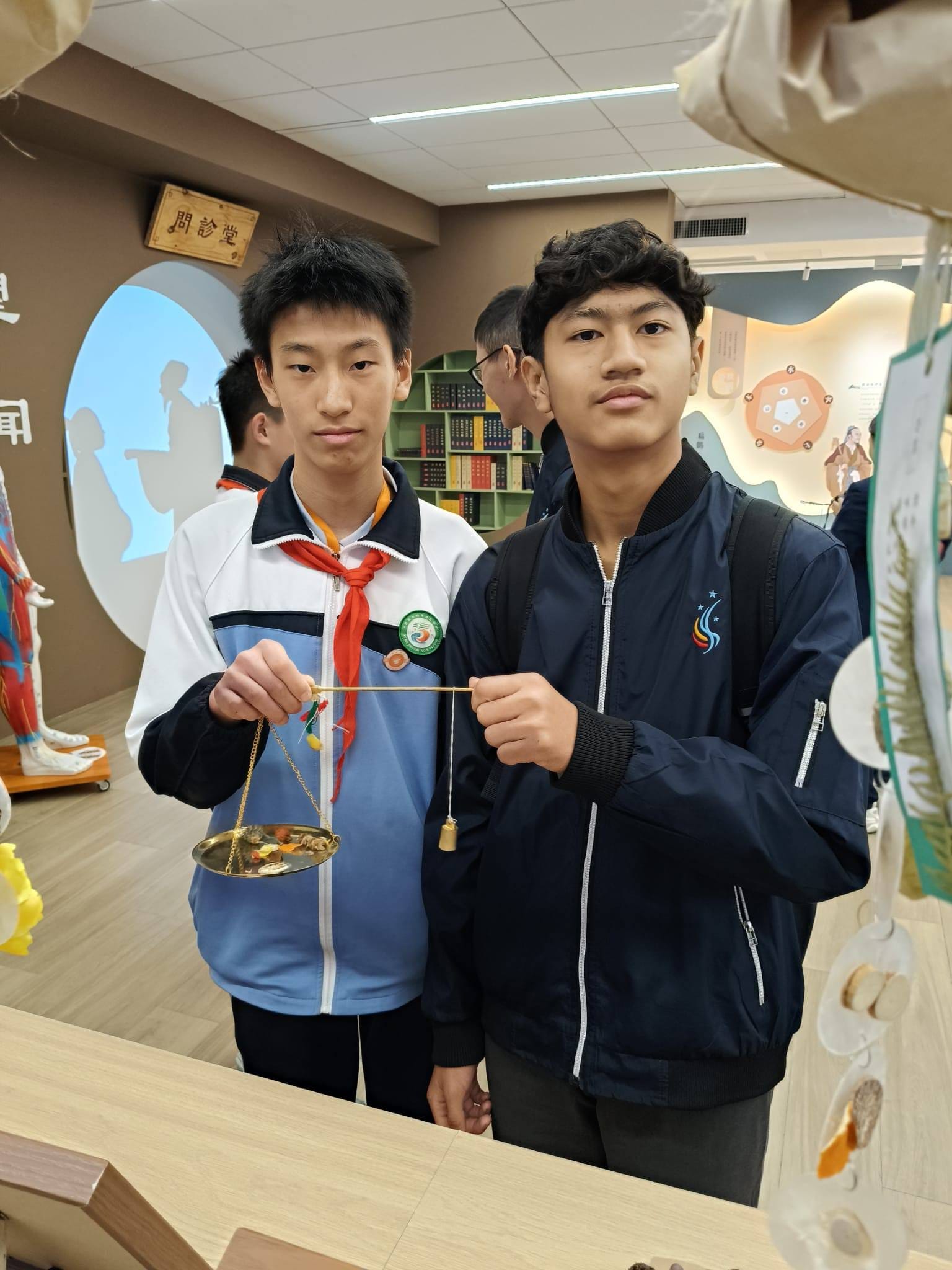 Two teenage boys hold a small traditional Chinese balance scale together inside a museum or education center.