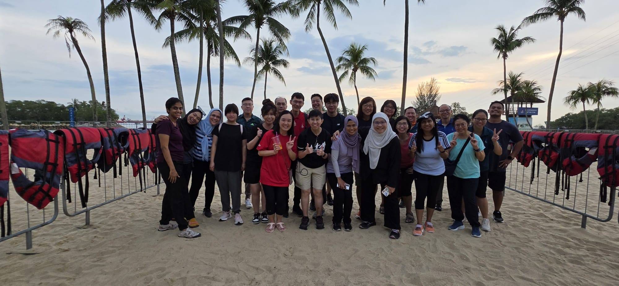 A large group of people posing on a sandy beach with red life jackets on railings and palm trees behind them.