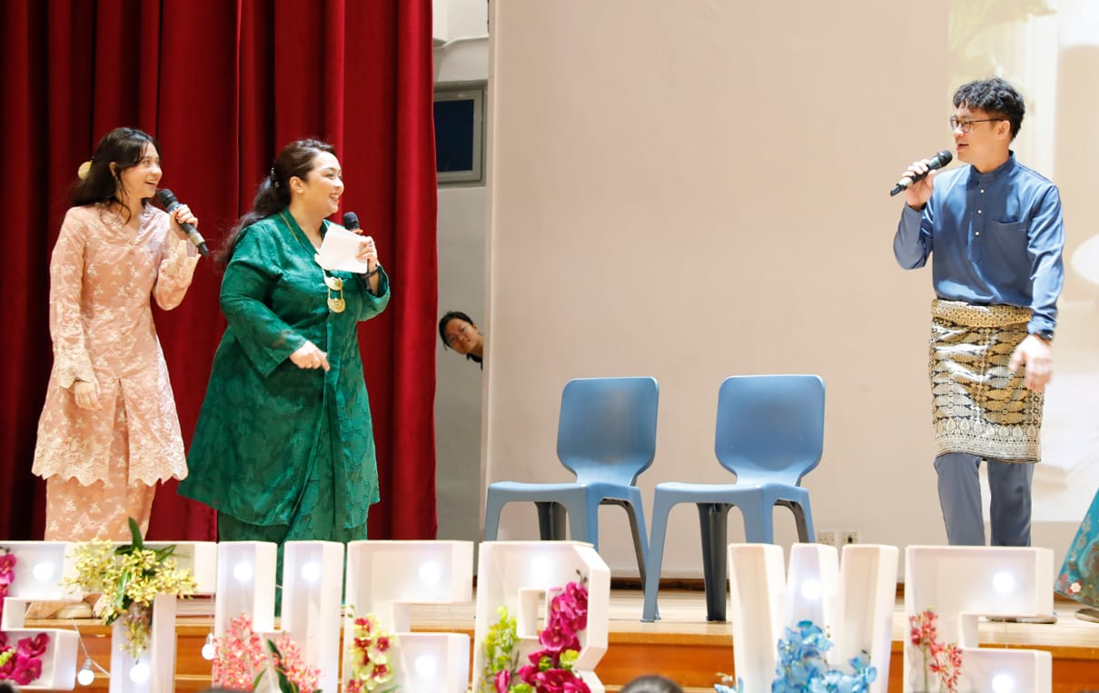 Three people in traditional attire hold microphones on a stage with red curtains, flowers, and blue chairs nearby.