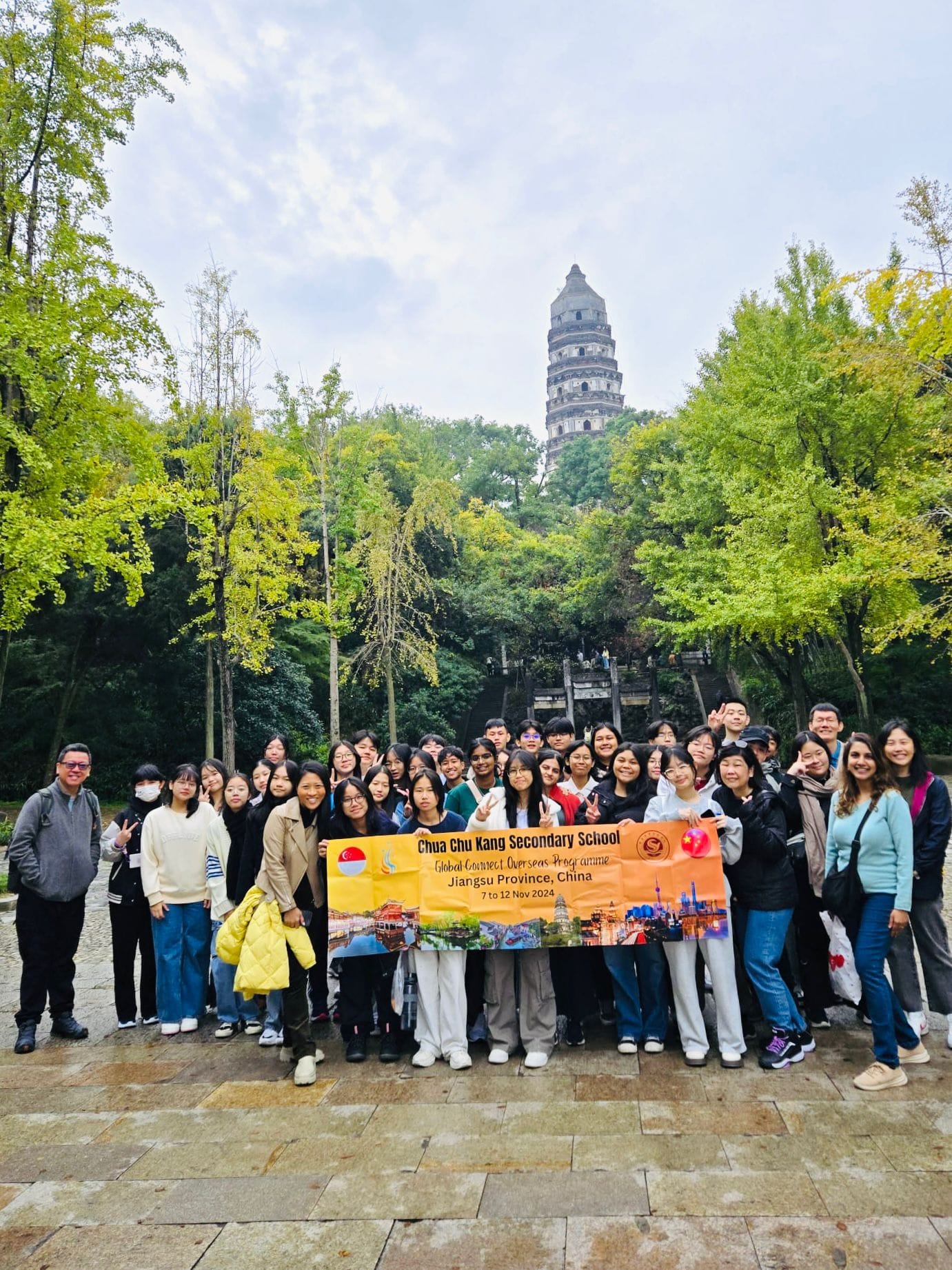A large student group holds a banner for Chua Chu Kang Secondary School's China trip, with a tall pagoda behind them.