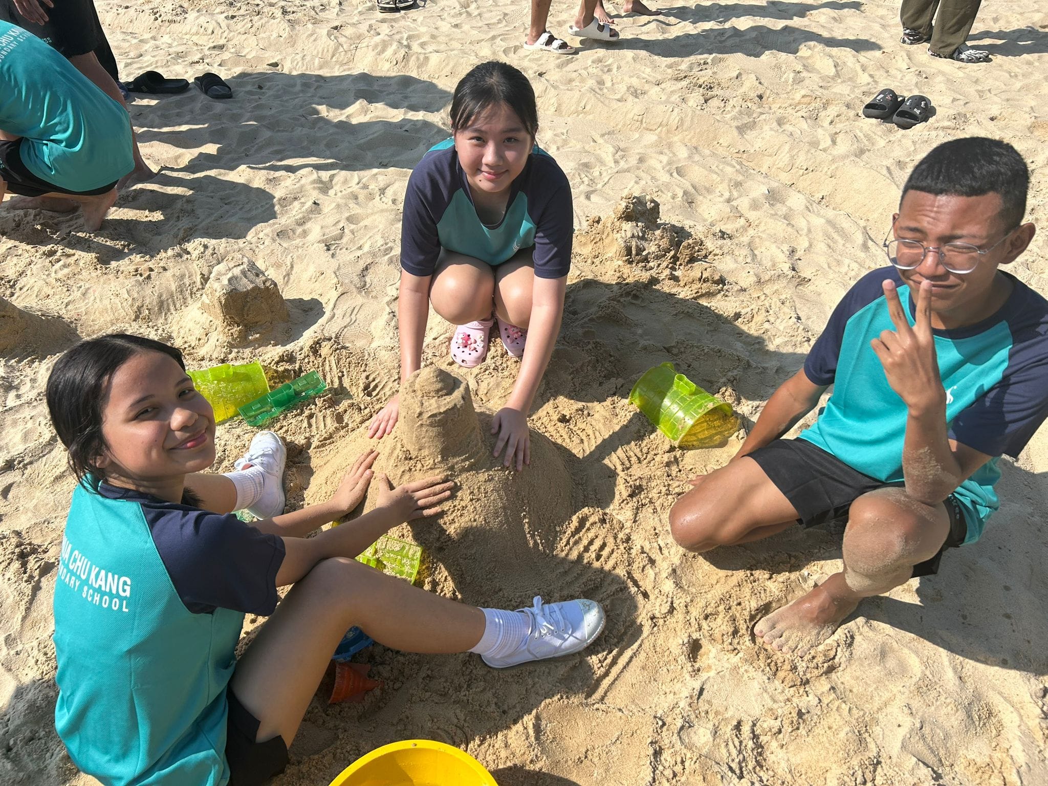 Three students in teal uniforms smile while building a sandcastle on a beach using green plastic molds.