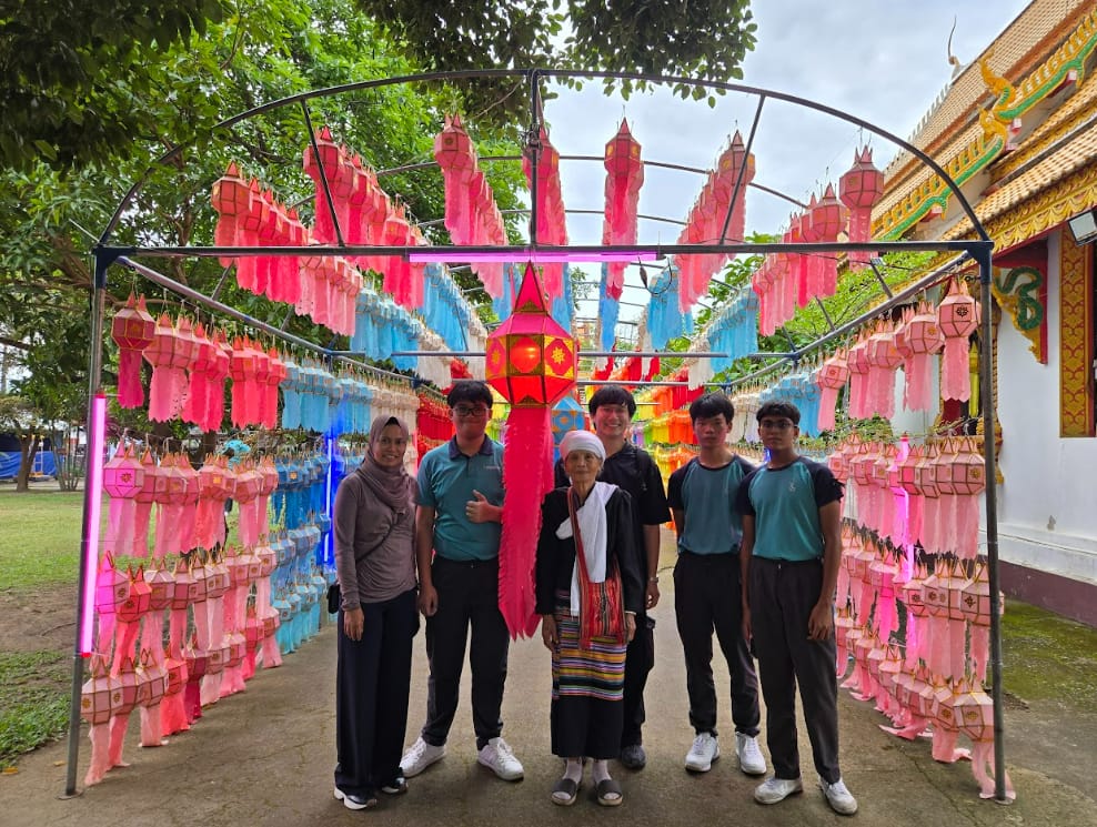 Six people stand under a metal arch covered with colorful pink and blue hanging lanterns near a temple.