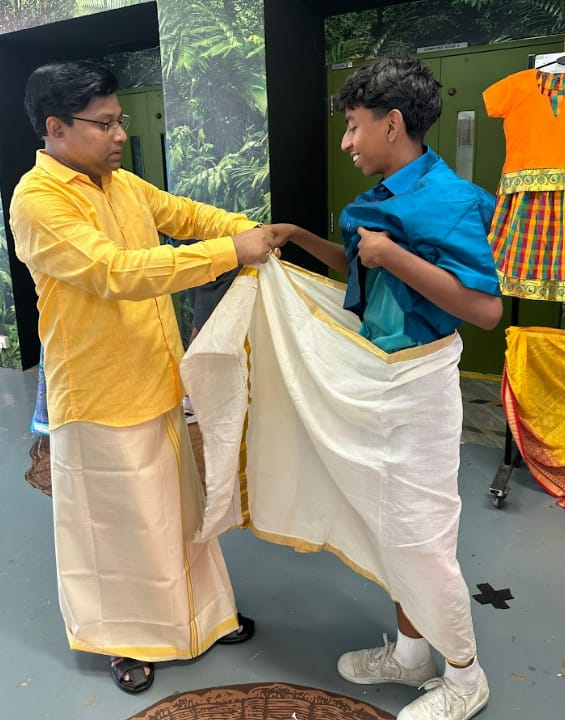 A man in a yellow kurta helps a smiling boy in a teal shirt drape a white and gold dhoti.