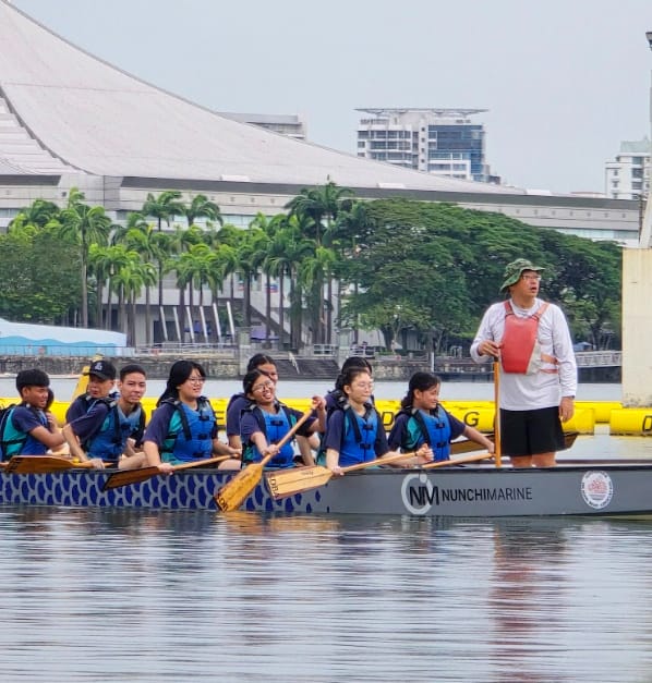 A team of young paddlers in life jackets row a Nunchi Marine dragon boat on a river with a city skyline behind.