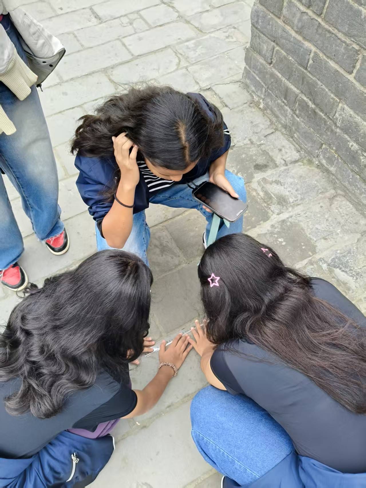 Three girls with dark hair huddle over a stone floor, one holding a phone, near a brick wall.