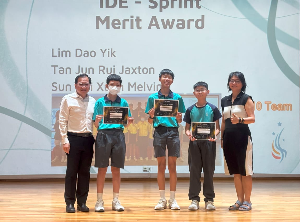 Three students hold certificates on stage for the IDE Sprint Merit Award, flanked by two adults.