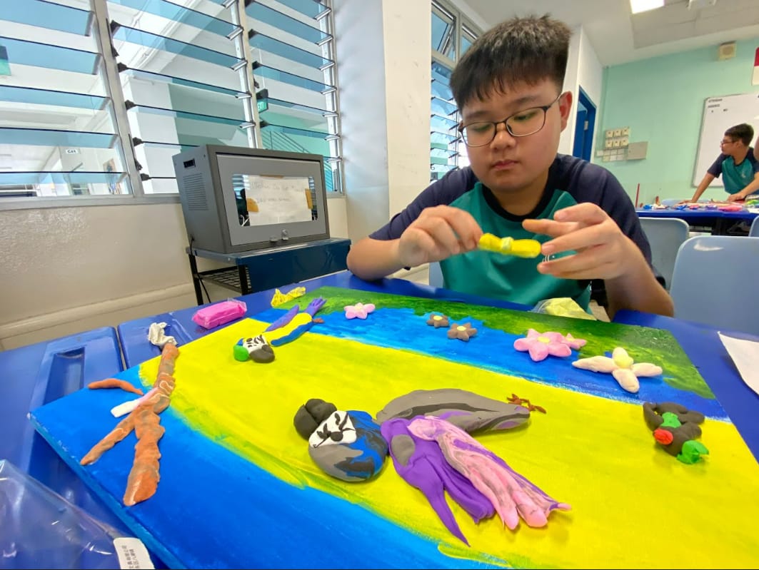 Boy with glasses sculpting colorful clay figures on a painted blue and yellow board in a classroom.