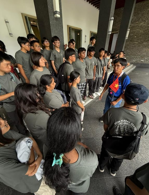 A large group of young people in matching gray shirts gathered around a guide holding a small flag indoors.
