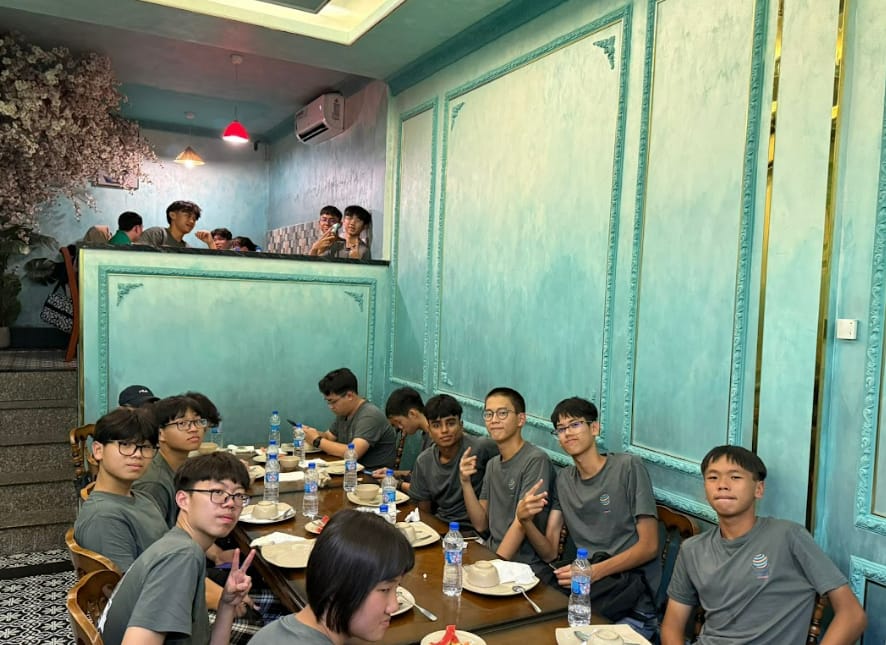 A group of young people in gray shirts sit at a dining table in a teal-walled restaurant, some making peace signs.