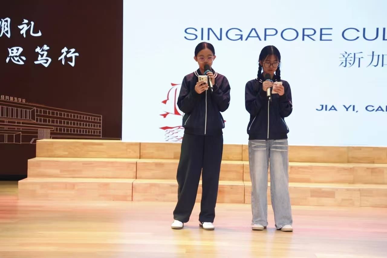 Two girls in matching jackets hold microphones and phones on a stage with a Singapore-themed backdrop.