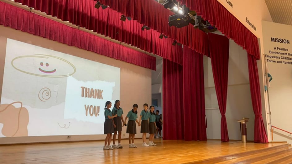 Five students in uniforms stand on a school stage holding hands beside a projected "Thank You" slide.