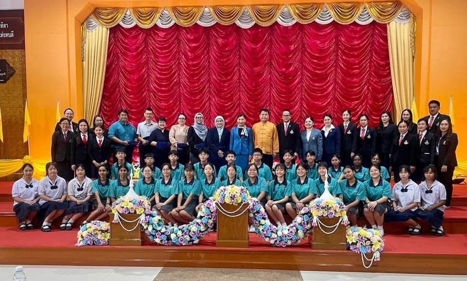 A large group of students and adults pose in front of a red and gold draped stage with colorful flower arrangements.