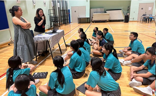 Two adults stand near a laptop table while students in teal shirts sit on a gymnasium floor listening.