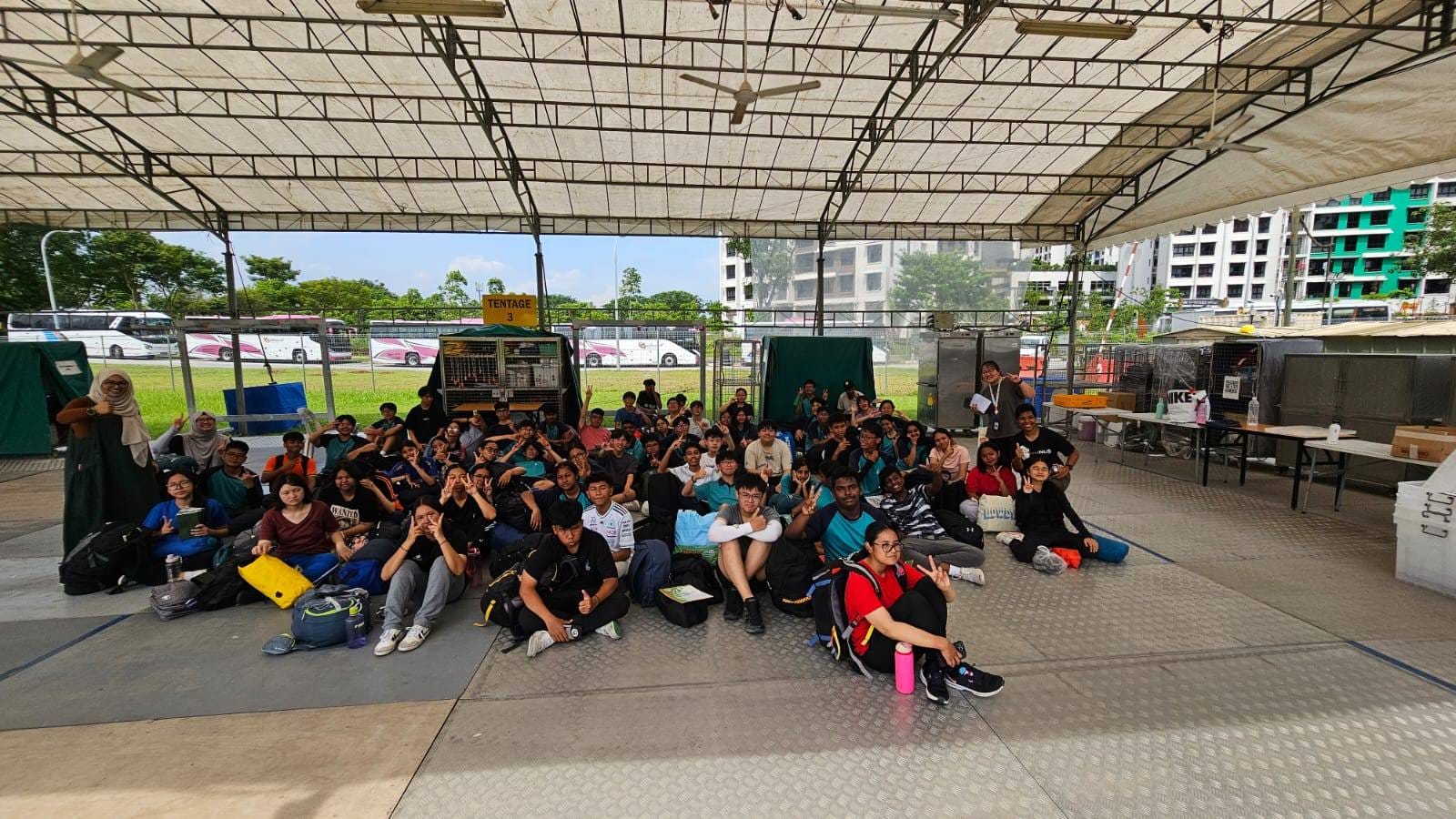 Large group of students sitting and posing under an outdoor tentage structure with buses and buildings in the background.