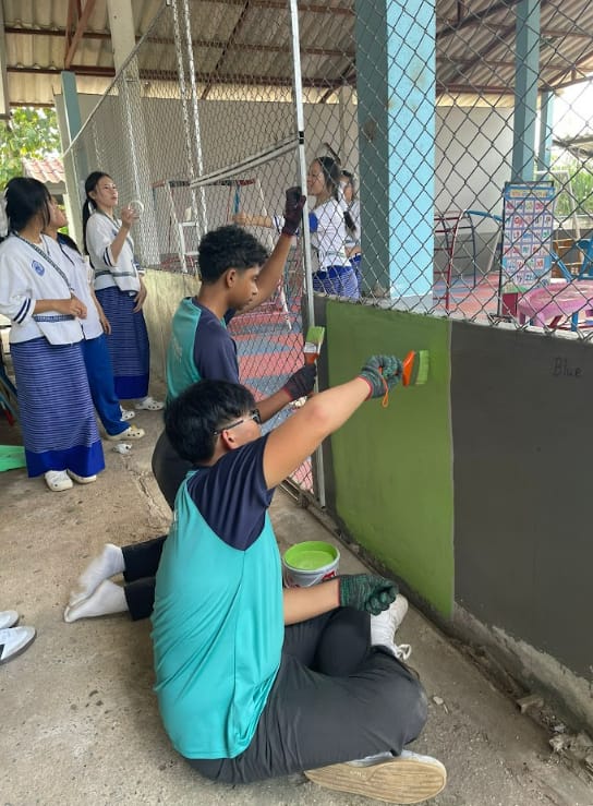 Two students painting a wall green with rollers while others watch nearby under a covered outdoor area.
