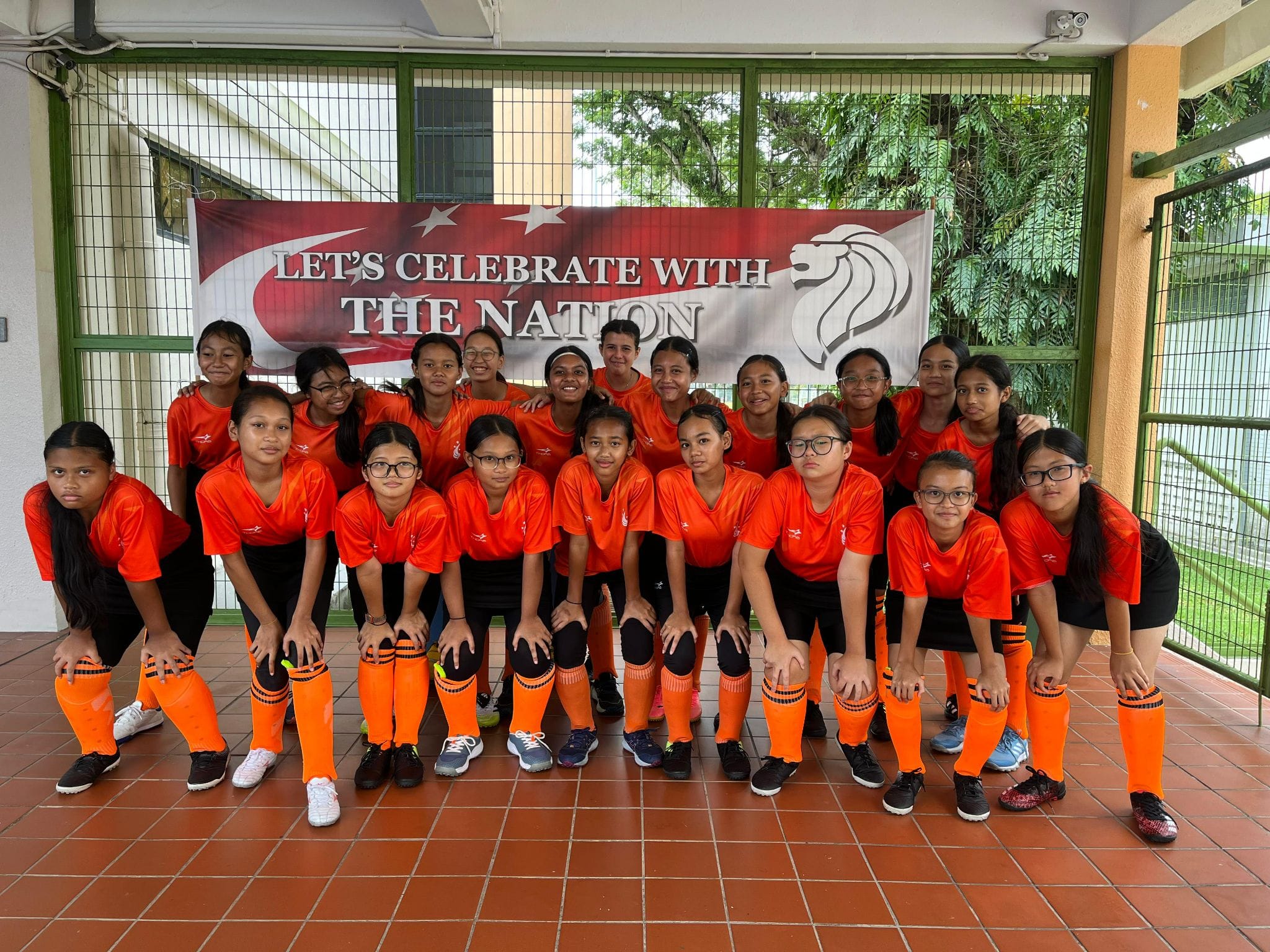 A group of girls in orange jerseys and socks pose in front of a banner reading "Let's Celebrate With The Nation."