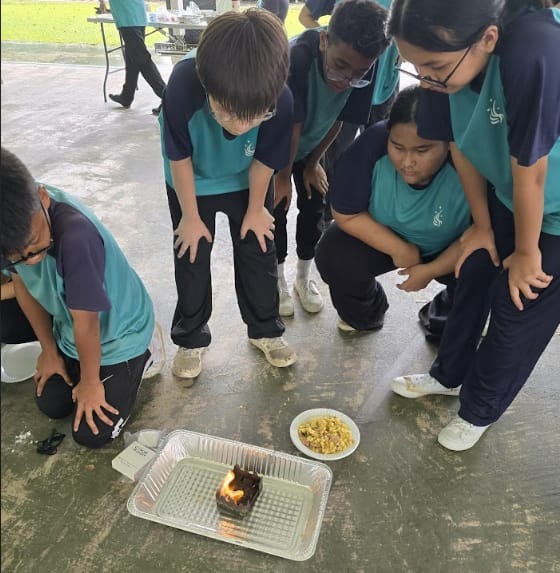 Students in teal uniforms crouch around a small flame burning in a foil tray on the ground.