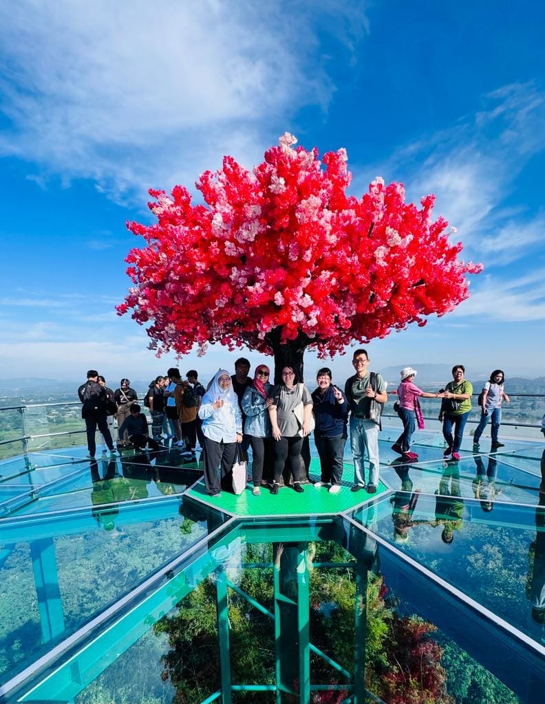 A group of people posing under a vibrant red flowering tree on a glass floor sky deck with mountain views.