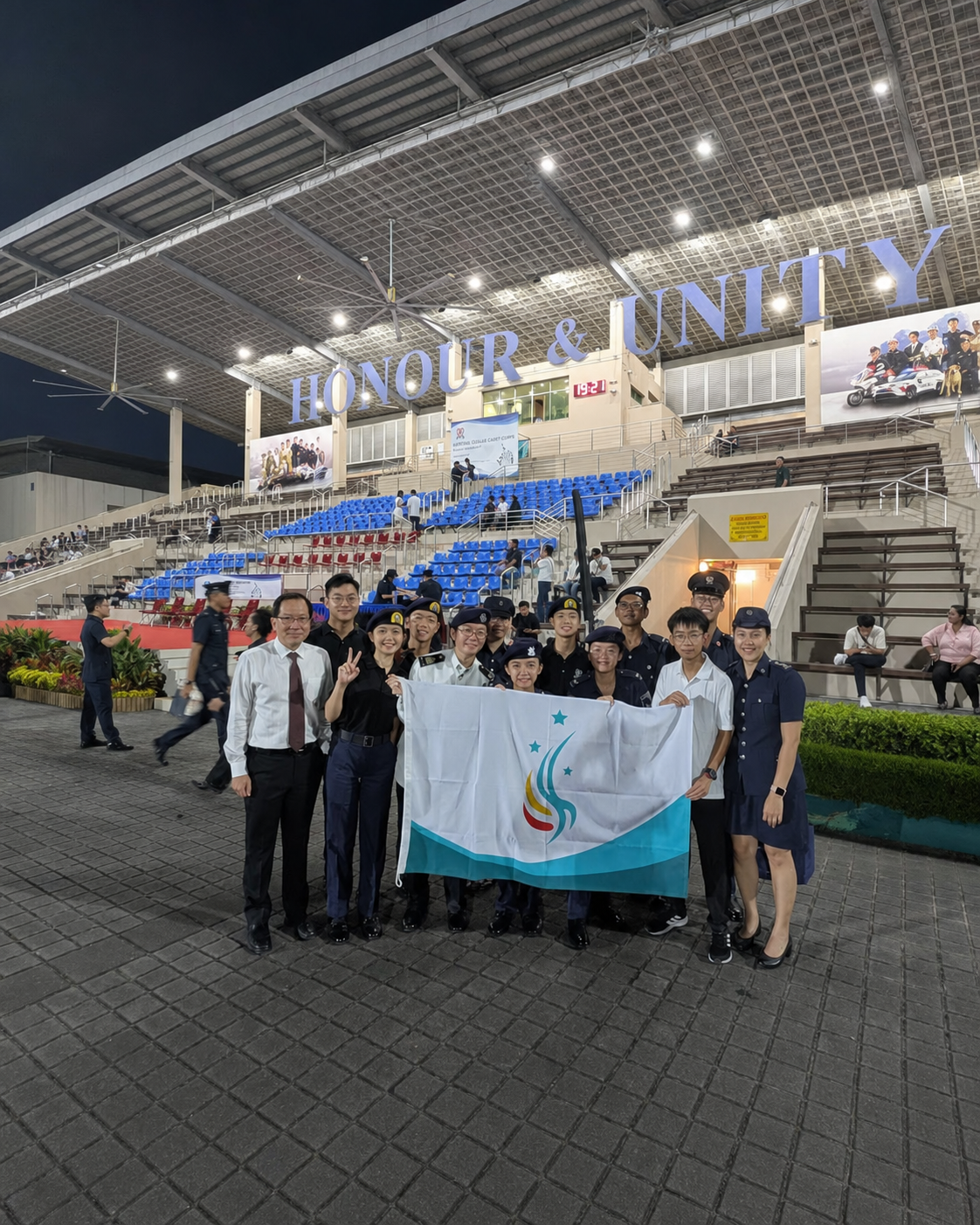 Group of uniformed cadets and adults posing with a flUnit Recognition Award (NPCC) Distinction (Awarded for 2024)ag inside a hall with "Honour & Unity" sign overhead.