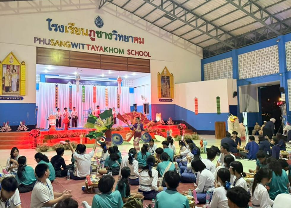 Students sit on the floor watching a colorful traditional Thai performance on a decorated stage at Phusangwittayakhom School.